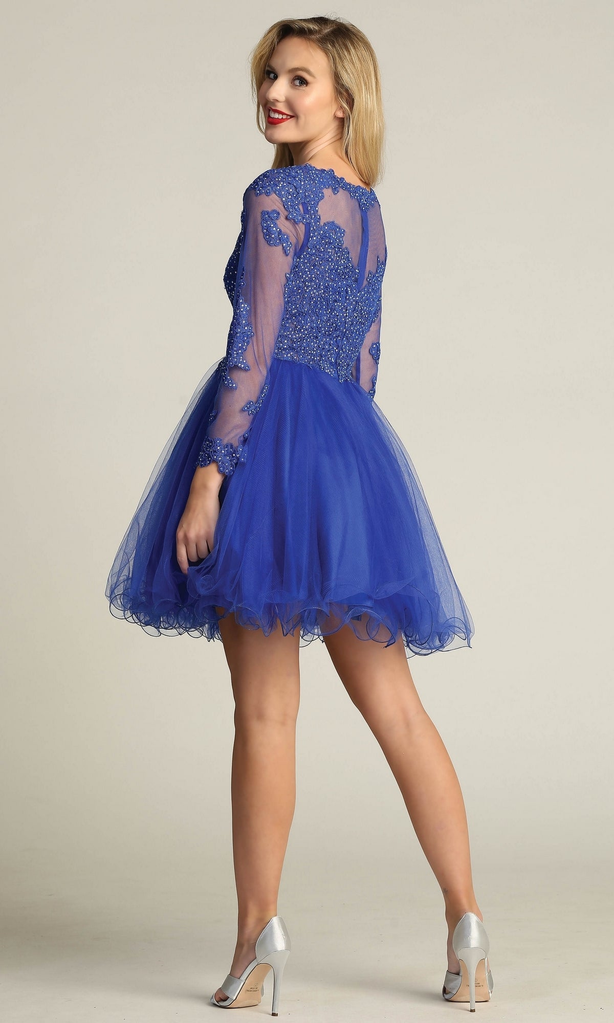 A woman poses and smiles in the Beaded Long Sleeve Short Homecoming Dress DM4003, a royal blue lace and tulle semi-formal dress with a beaded bodice, paired with silver high heels against a plain background.